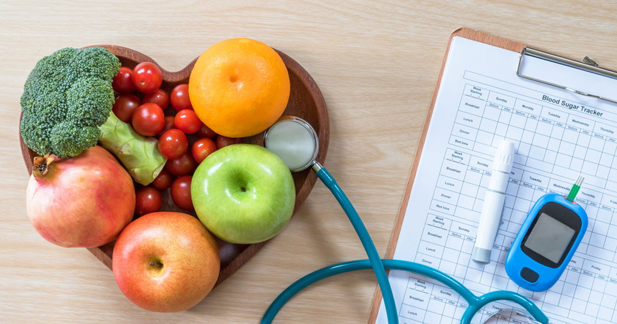 Heart shaped bowl of fruit and vegtables near a blood sugar monitor, stethescope, and blood sugar chart