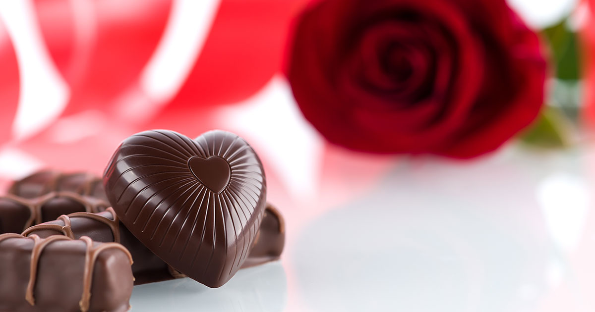 A chocolate heart rests against other chocolate candy in the foreground with a blurry rose sitting in the background.
